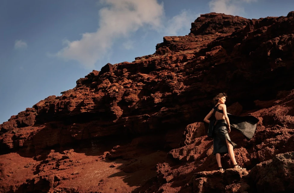 Woman in black dress posing on a red rock hill under a blue sky with clouds, back view, fashion editorial photography.