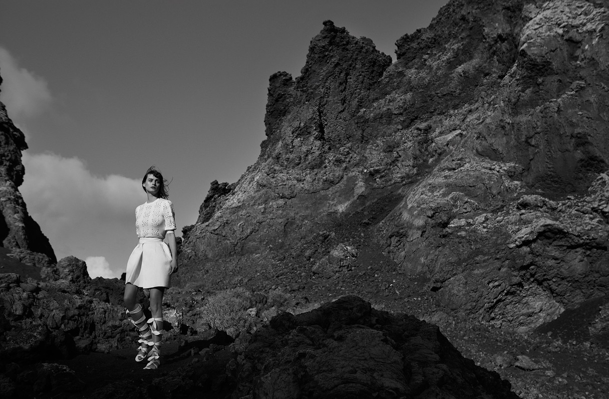 Black and white portrait of a woman standing against a rocky mountain landscape wearing patterned top and skirt.