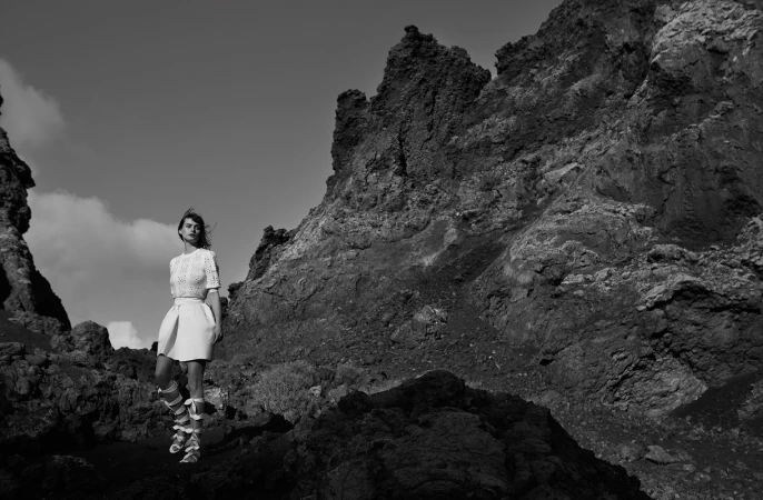 Black and white portrait of a woman standing against a rocky mountain landscape wearing patterned top and skirt.