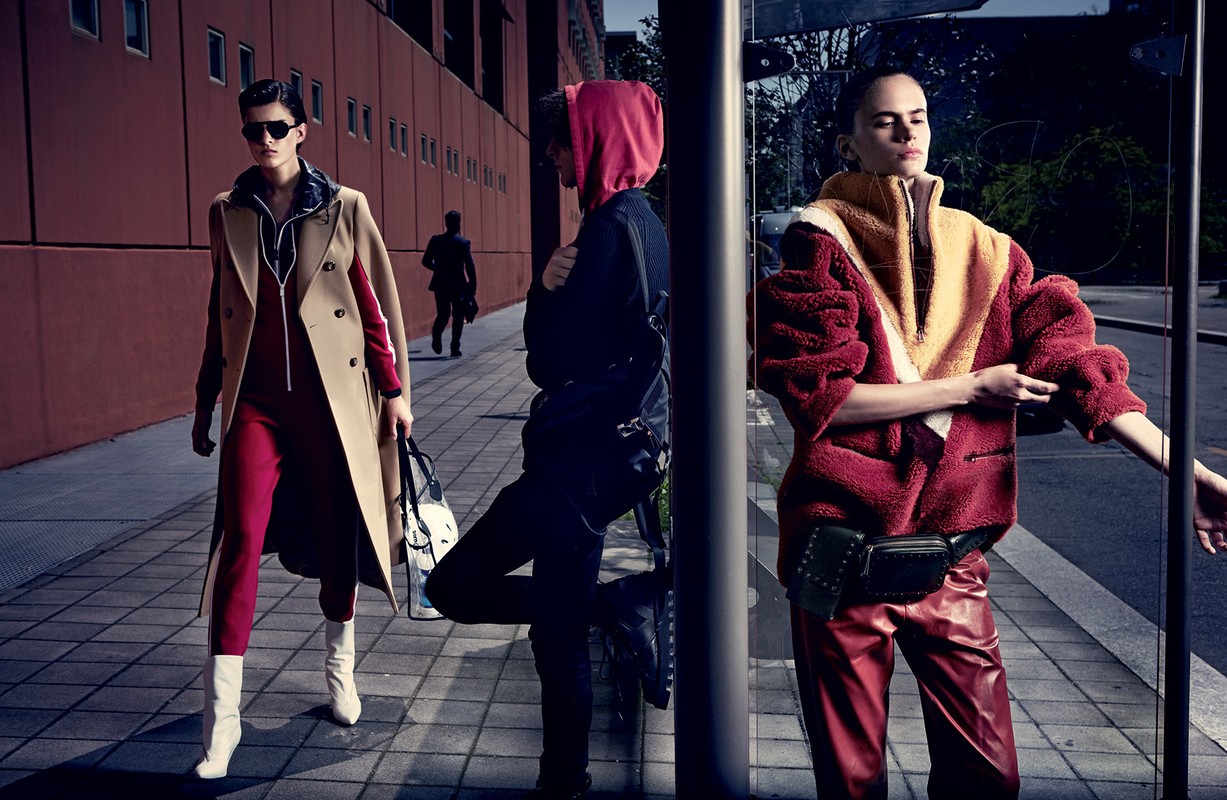 Three models pose in modern urban fashion shoot: woman in tan coat, red jumpsuit, white boots; another in red hoodie and leather pants.