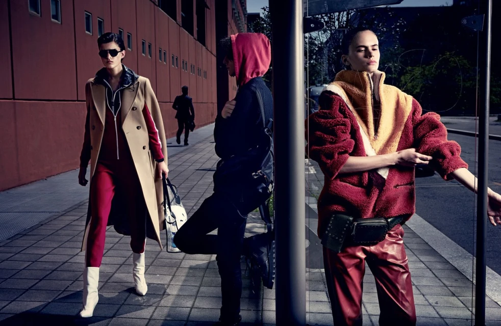 Three models pose in modern urban fashion shoot: woman in tan coat, red jumpsuit, white boots; another in red hoodie and leather pants.