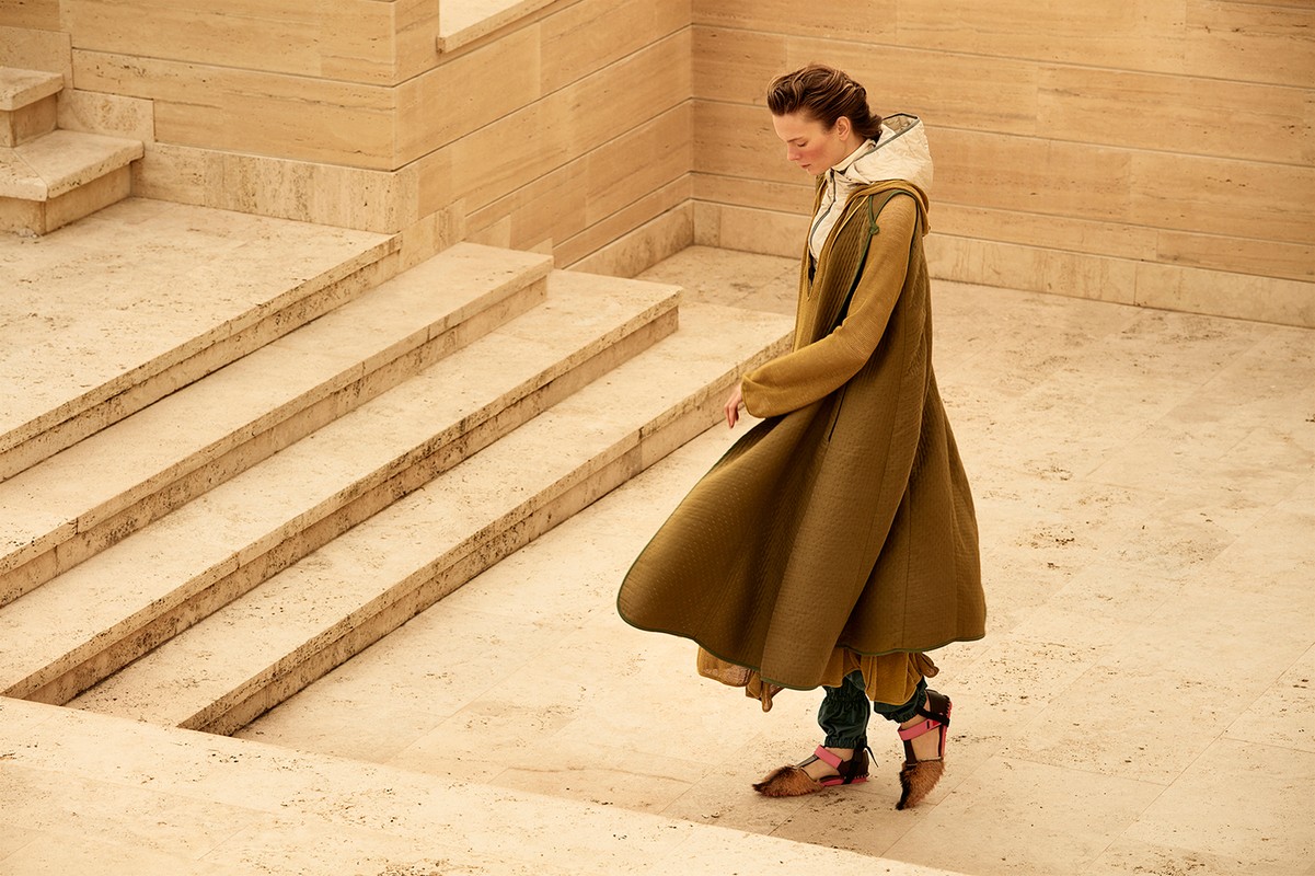 A woman in a long, olive-green coat descends a light-tan stone staircase in an elegant outdoor space with dappled sunlight.