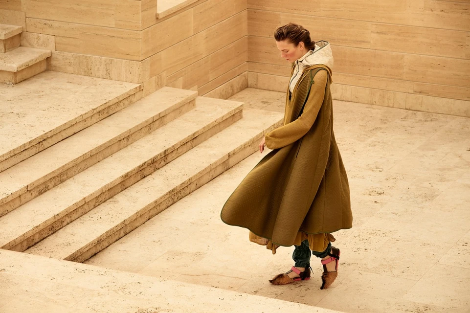 A woman in a long, olive-green coat descends a light-tan stone staircase in an elegant outdoor space with dappled sunlight.