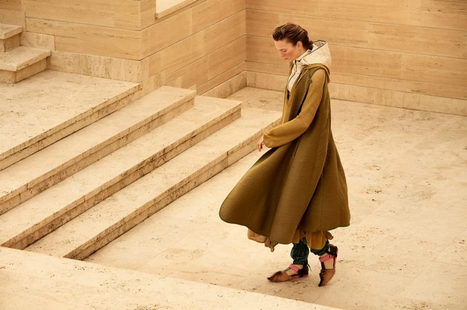 A woman in a long, olive-green coat descends a light-tan stone staircase in an elegant outdoor space with dappled sunlight.