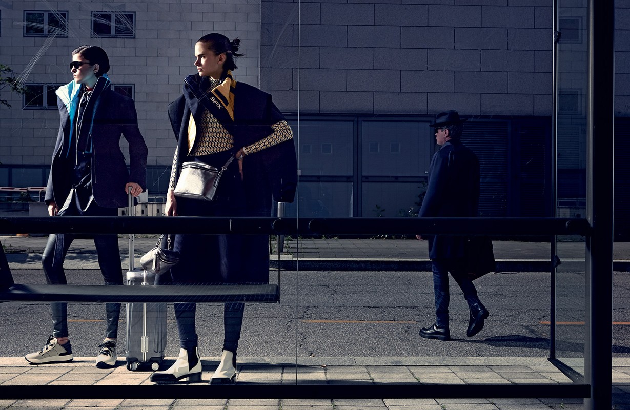 Two female models posing at a glass bus shelter with a rolling suitcase, photographed by Takay in harsh sunlight.