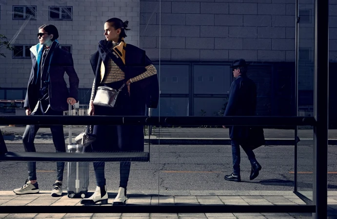 Two female models posing at a glass bus shelter with a rolling suitcase, photographed by Takay in harsh sunlight.
