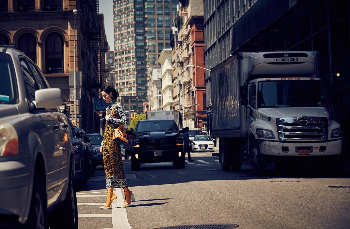 Woman in patterned jumpsuit crosses city street with cars and truck on sunny day. Buildings line the urban landscape in background.