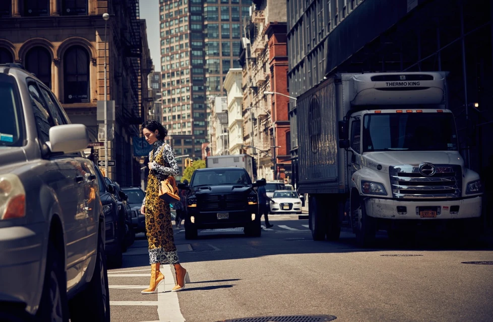 Woman in patterned jumpsuit crosses city street with cars and truck on sunny day. Buildings line the urban landscape in background.