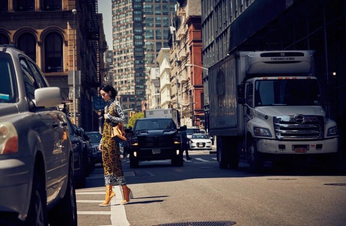 Woman in patterned jumpsuit crosses city street with cars and truck on sunny day. Buildings line the urban landscape in background.
