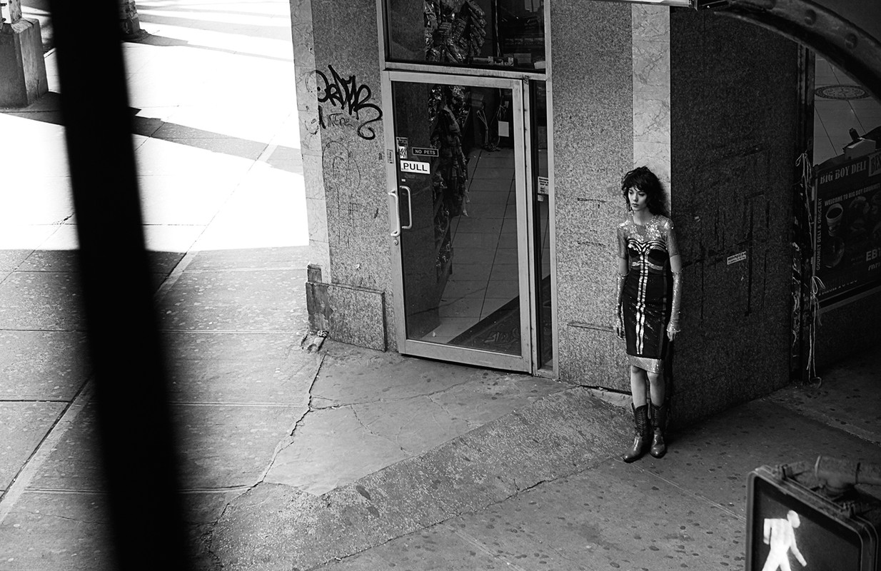 Woman in fitted black dress stands against a building on a city sidewalk, next to a door with graffiti. Black and white photography.