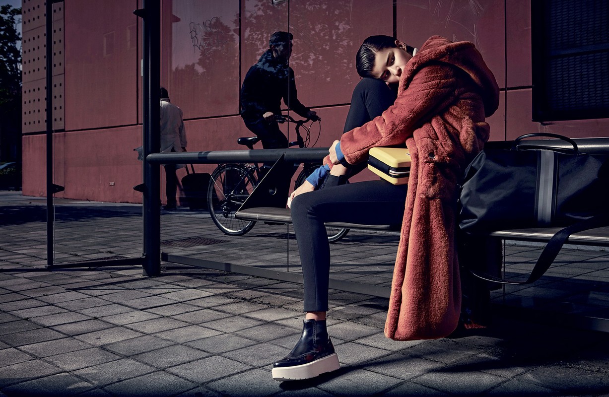 Fashion photography: A young woman in a warm coat sits at a bus stop next to a travel bag, with a cyclist and pedestrians in the backdrop.