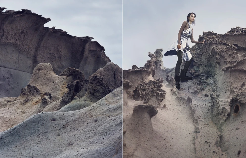 Fashion editorial photo of a model in a silver dress posing on a rugged, rocky coastline under an overcast sky with dramatic rock formations.