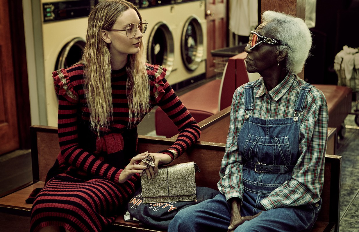 Two women, one in red/black stripes and the other in overalls, sit in a vintage laundromat with washing machines behind them.
