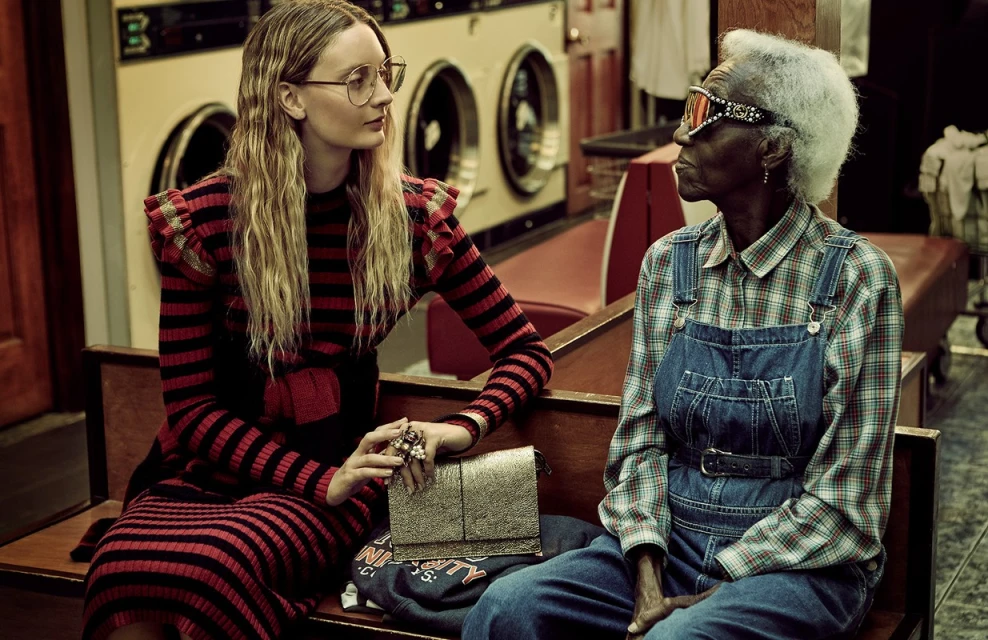 Two women, one in red/black stripes and the other in overalls, sit in a vintage laundromat with washing machines behind them.