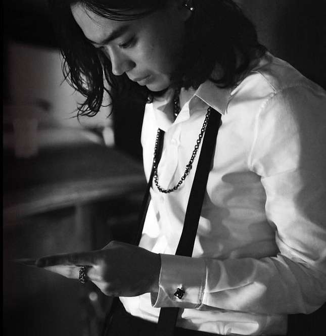 Monochrome portrait of an adult male with long hair, wearing a white shirt and suspenders, looking down and pointing at something.