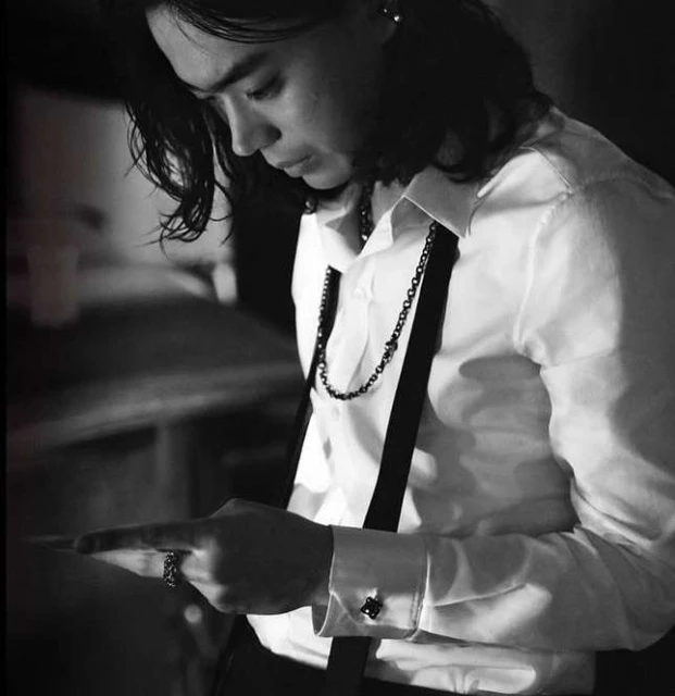 Monochrome portrait of an adult male with long hair, wearing a white shirt and suspenders, looking down and pointing at something.