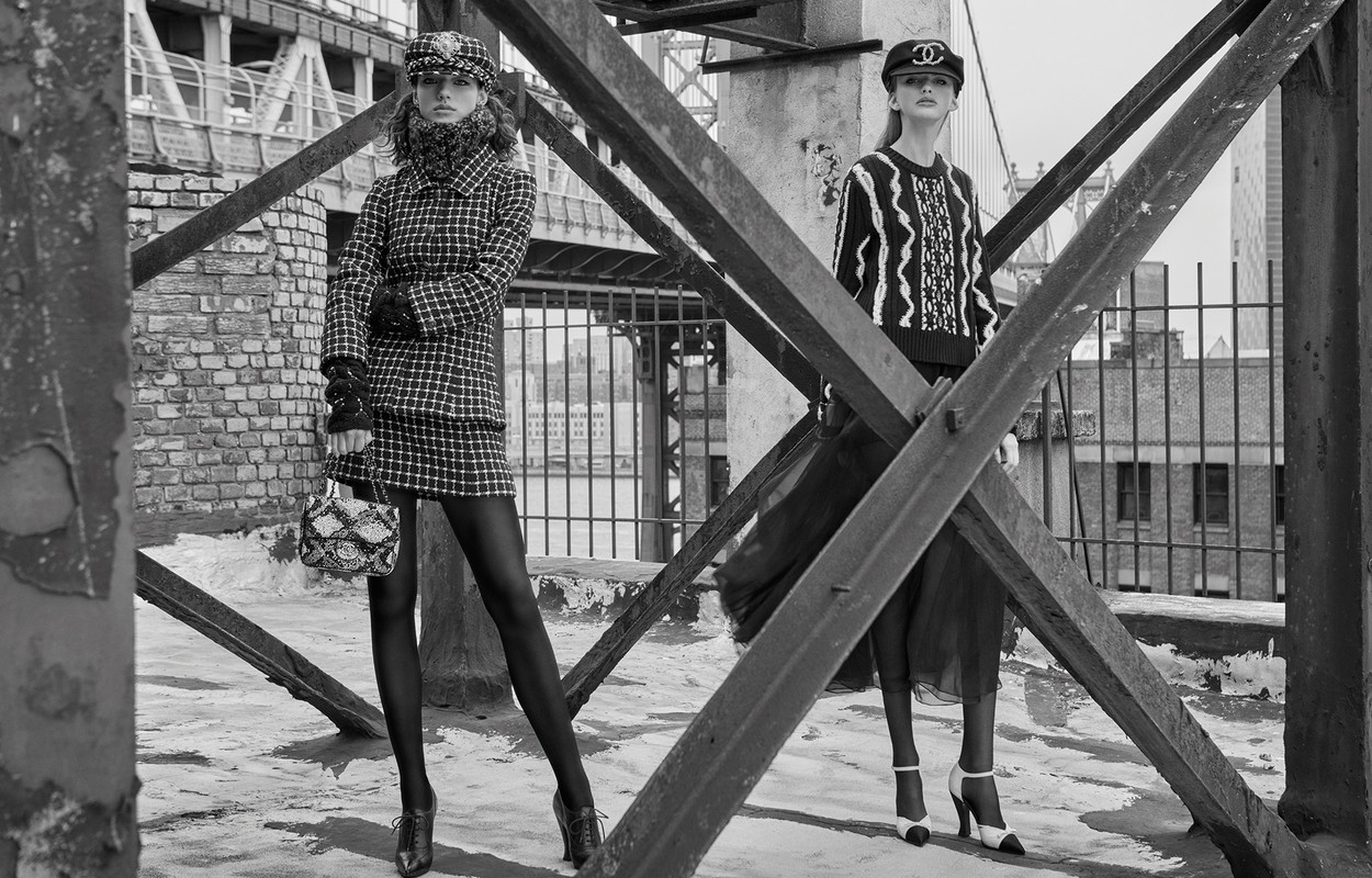 Two female models in tweed and knitwear posing on a rooftop behind structural bridge beams in a black and white editorial photograph.