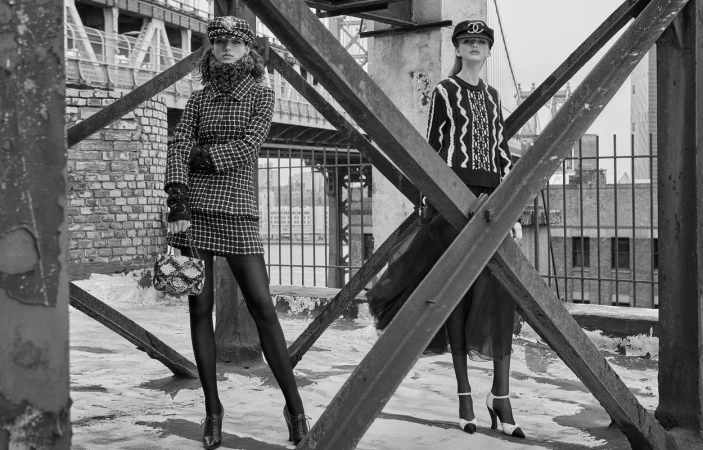 Two female models in tweed and knitwear posing on a rooftop behind structural bridge beams in a black and white editorial photograph.