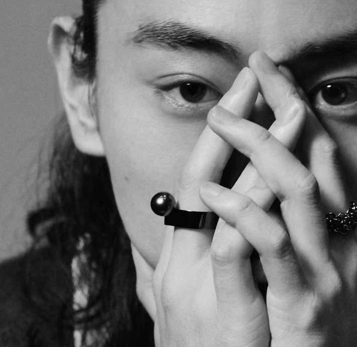 Close-up black-and-white studio shot of a person with long dark hair, hands up to the face, and a black ring with a sphere on it.