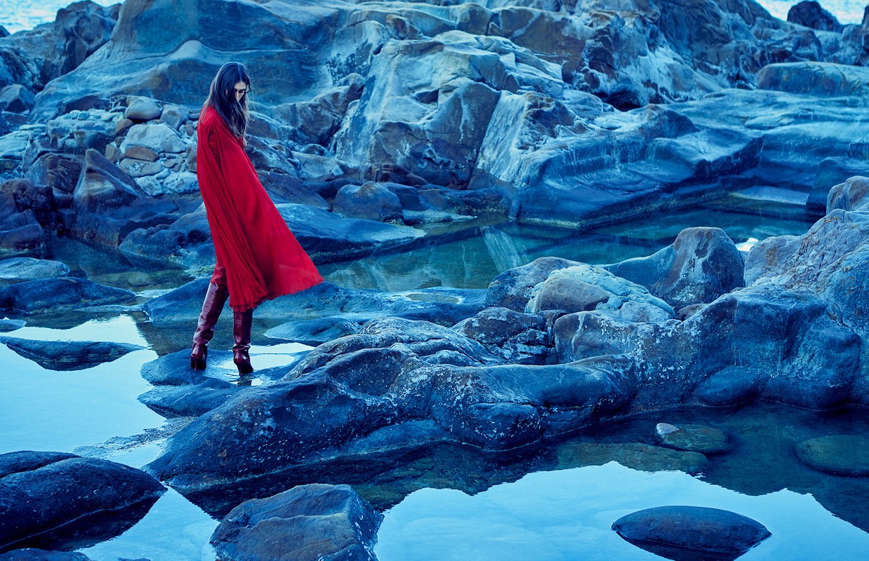 Woman in a red cloak walks on rocks by tranquil blue water. Moody, picturesque landscape with stone textures and reflective surface.