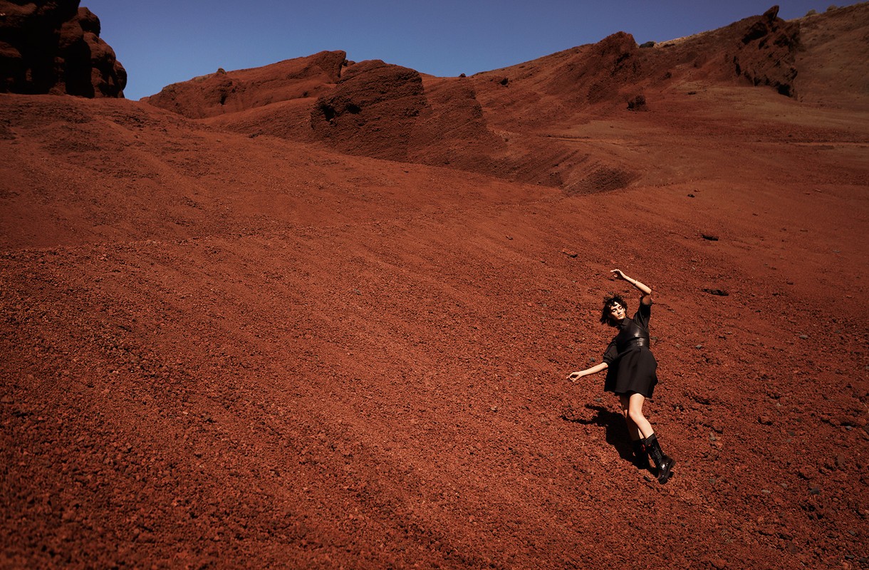 Woman in black dress poses on reddish-brown rocky hill under blue sky. Wide shot of outdoor fashion photography.
