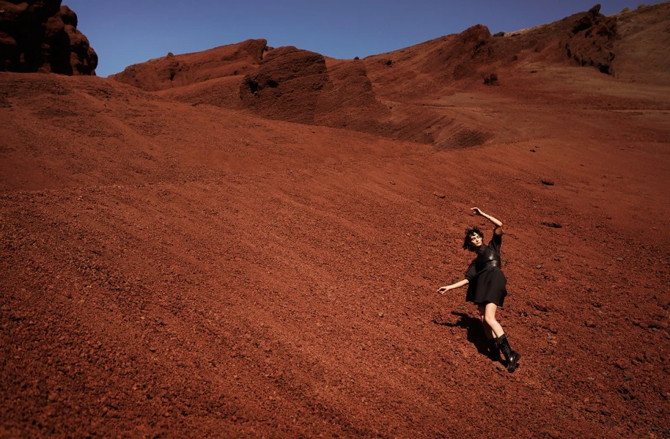 Woman in black dress poses on reddish-brown rocky hill under blue sky. Wide shot of outdoor fashion photography.