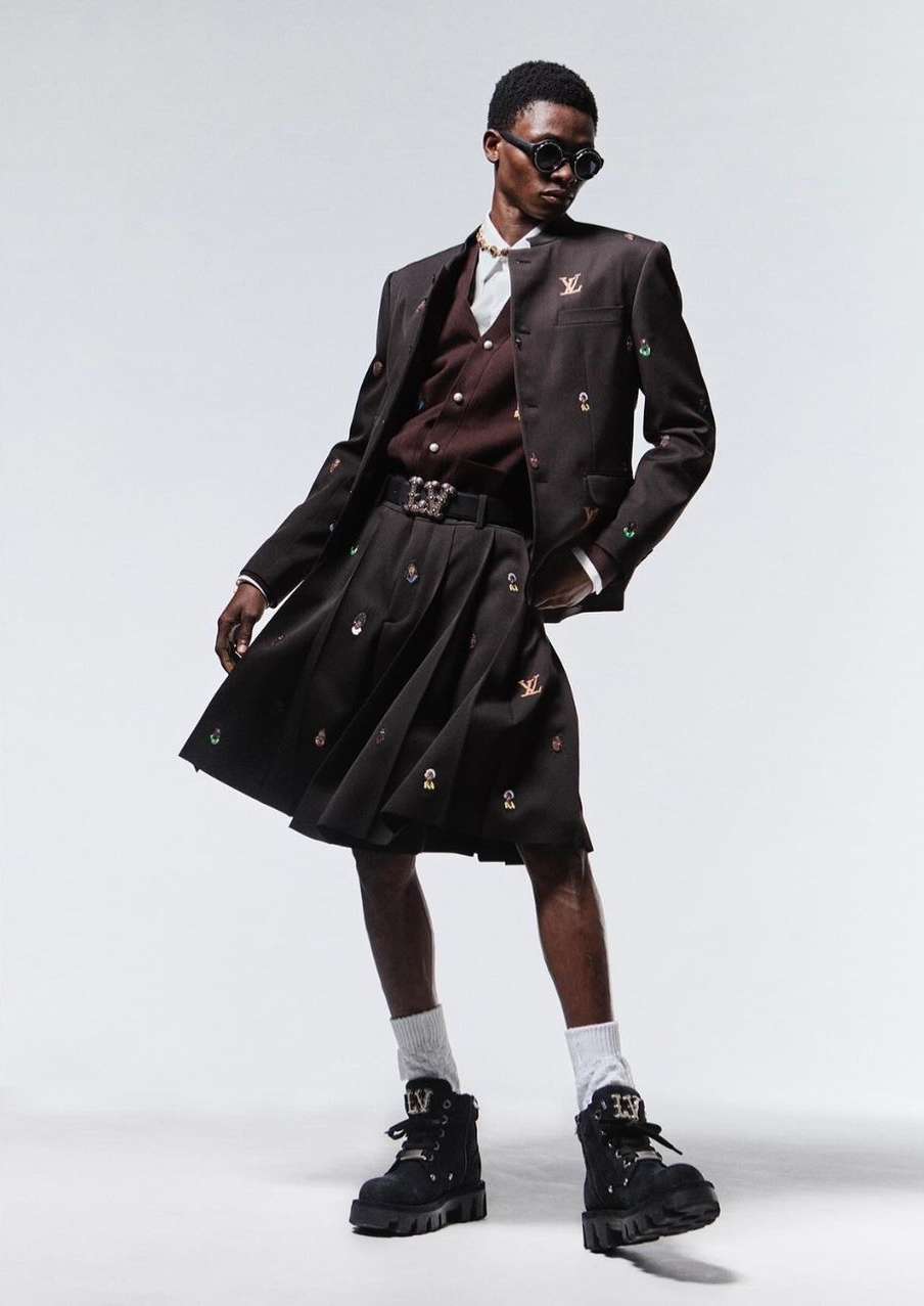 Fashionable adult male model in sunglasses, brown suit jacket, pleated kilt, white socks, and black boots posing in a photo studio.