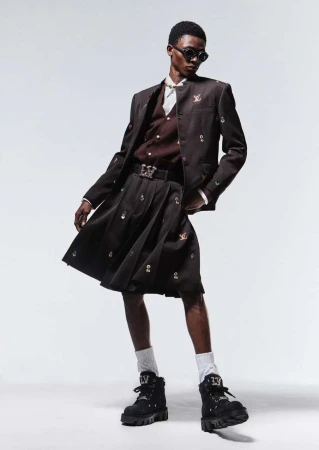 Fashionable adult male model in sunglasses, brown suit jacket, pleated kilt, white socks, and black boots posing in a photo studio.