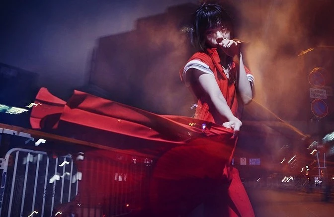 Singer in a red outfit performs on stage with motion blur and soft lighting, captured in a medium shot on an urban street at night.