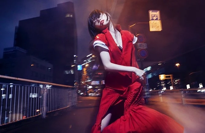Woman in a vibrant red dress posing dynamically on a city street at night under streetlights, photographed by Takay.