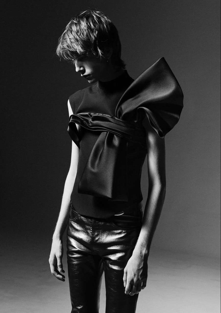 Black and white studio portrait of a person in a black sleeveless top with an oversized bow and shiny black pants.