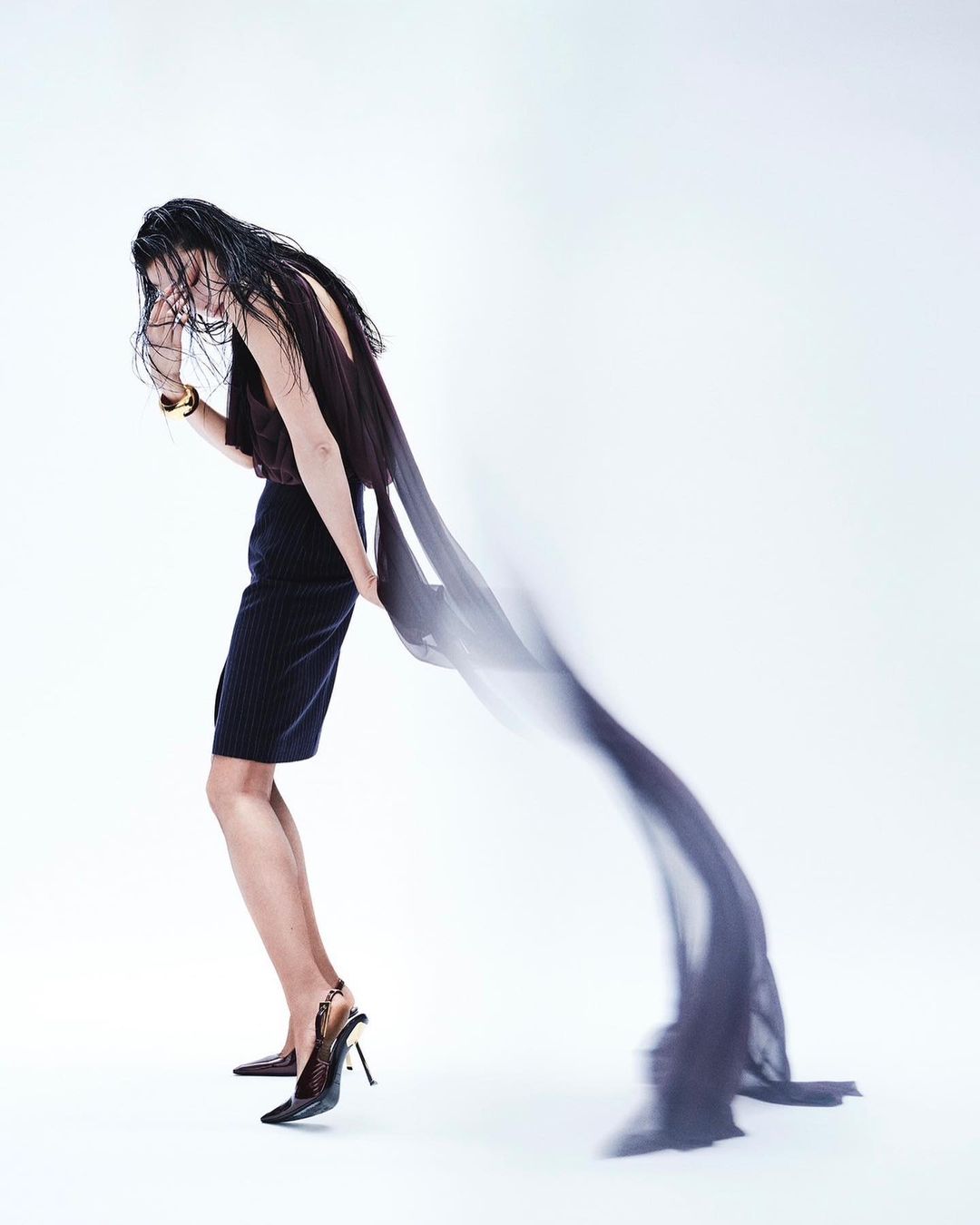 Fashion shot of a person wearing a dark top and skirt with heels, holding a long, semi-transparent scarf against a white backdrop.
