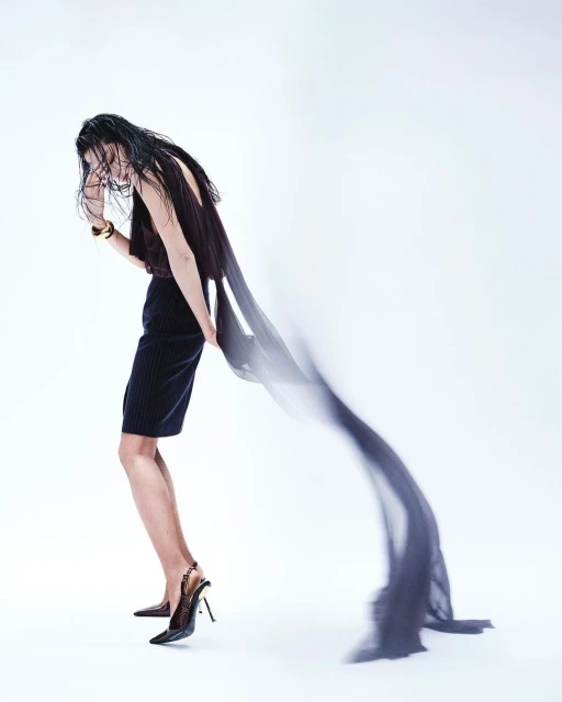 Fashion shot of a person wearing a dark top and skirt with heels, holding a long, semi-transparent scarf against a white backdrop.