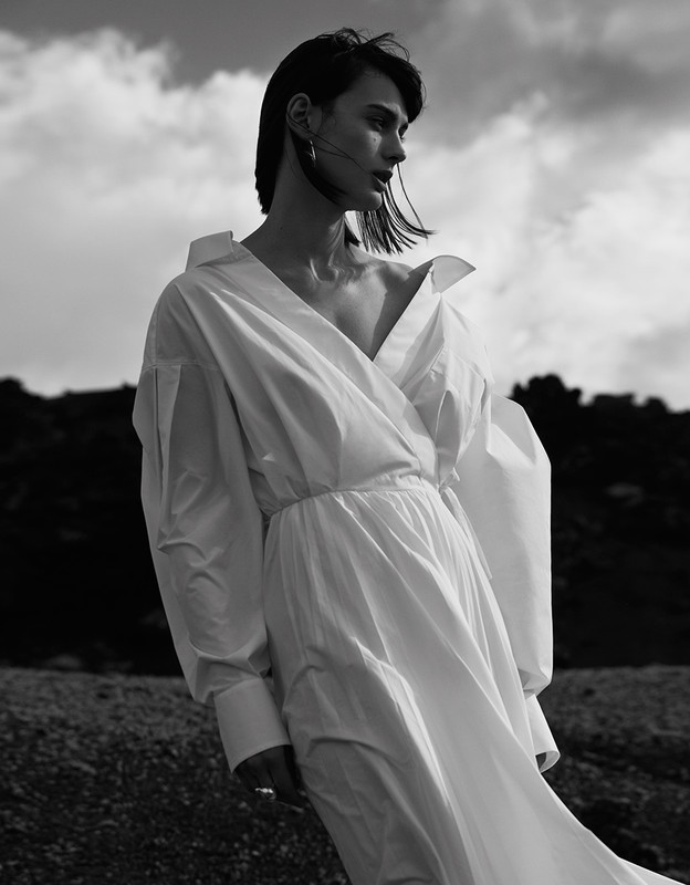 Monochrome portrait of a stylish adult female in white dress, wind blowing, standing outdoors with dramatic sky in the background.