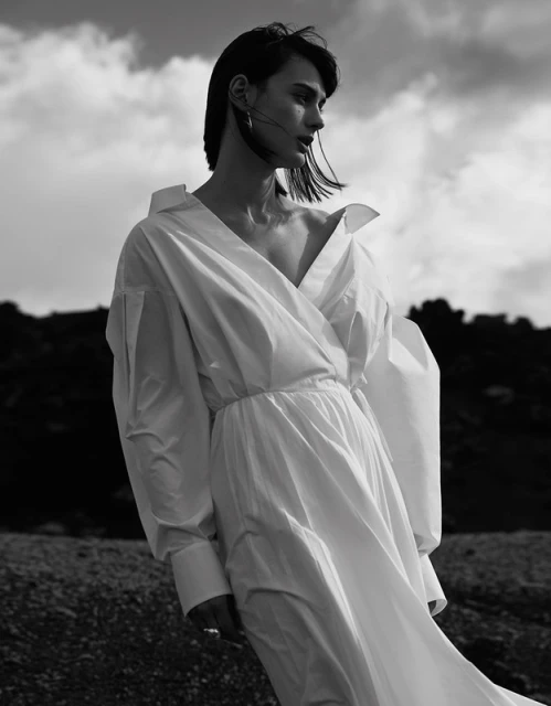 Monochrome portrait of a stylish adult female in white dress, wind blowing, standing outdoors with dramatic sky in the background.