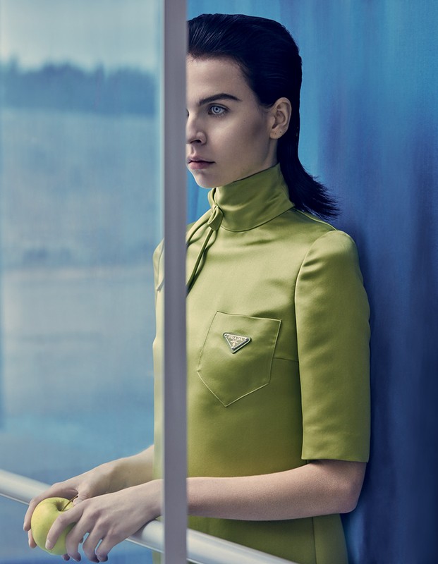 A woman in a green dress stands by a blue wall, holding a green apple. The Prada logo is on the dress's pocket.