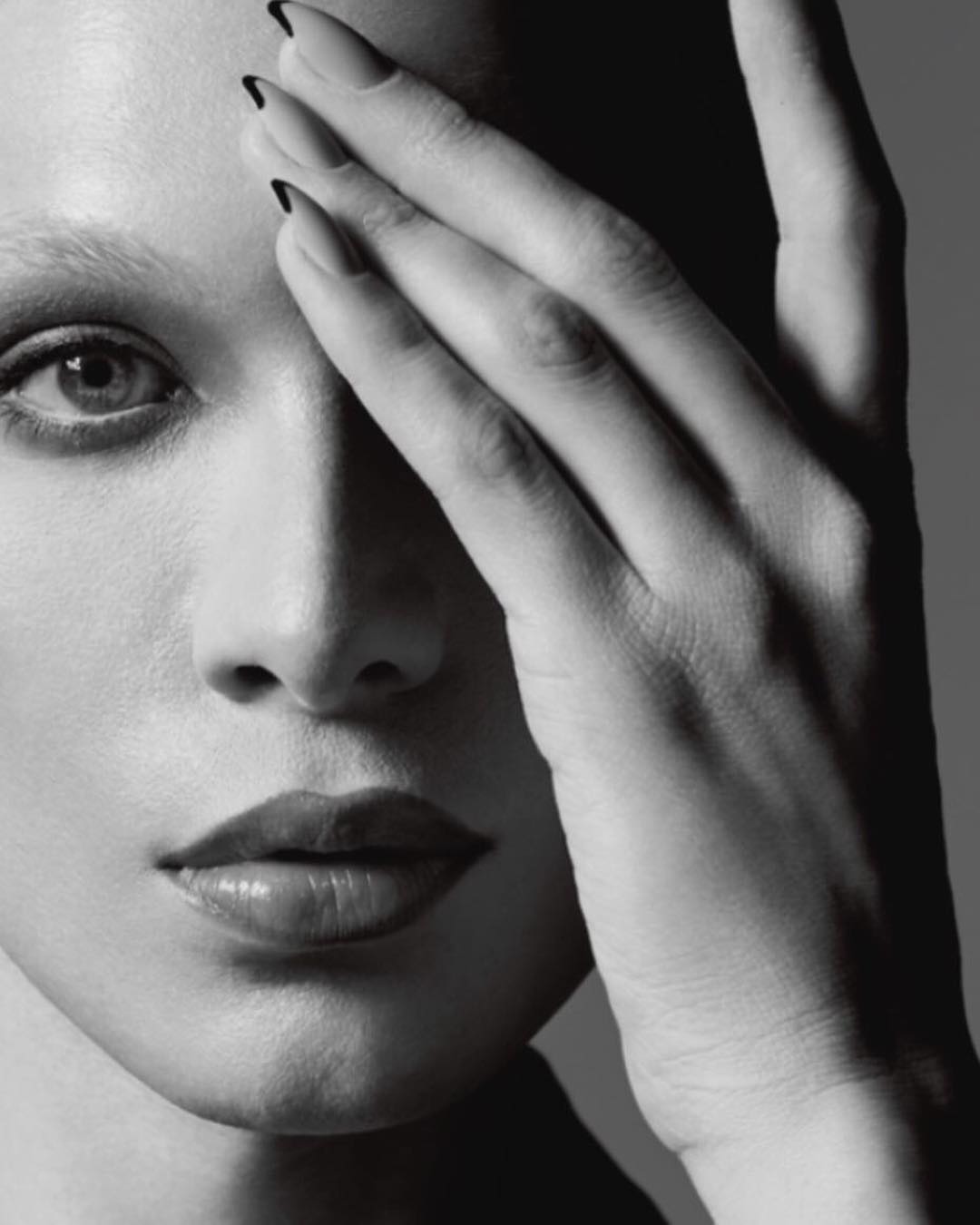 Monochrome portrait of a woman with hand covering part of her face, showcasing her glossy lips and manicured nails.