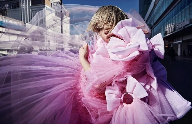 Model wearing a voluminous pink tulle dress with large bows outdoors against an urban street background, photographed by Takay.