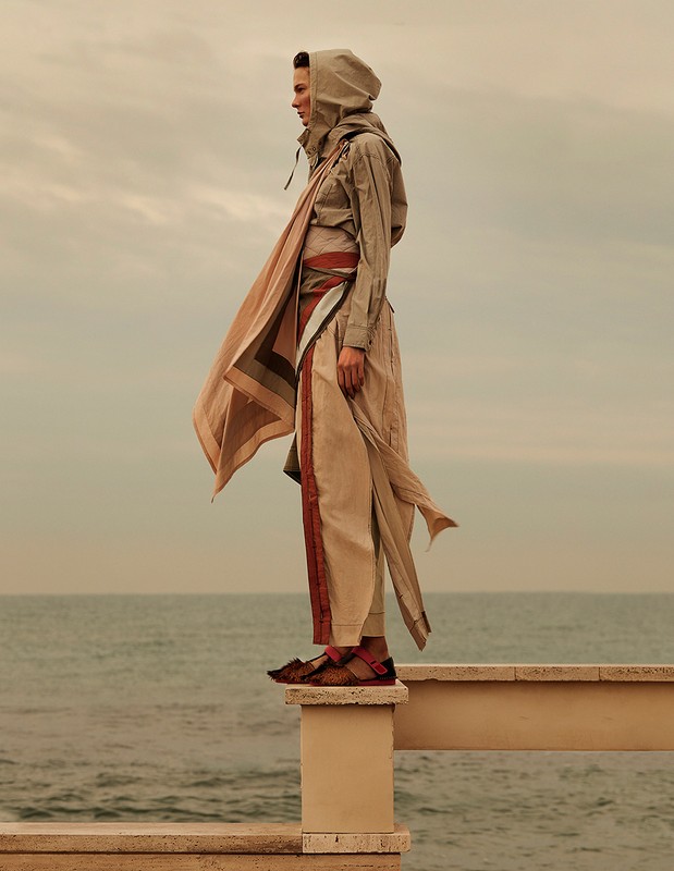 Fashion model stands on a ledge at the beach wearing a hooded earth-toned outfit, with ocean and cloudy sky visible in the background.