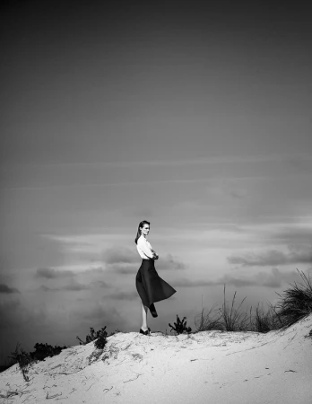 Model standing on a sandy hill with arms crossed against a cloudy sky in a monochrome fashion photograph by Takay