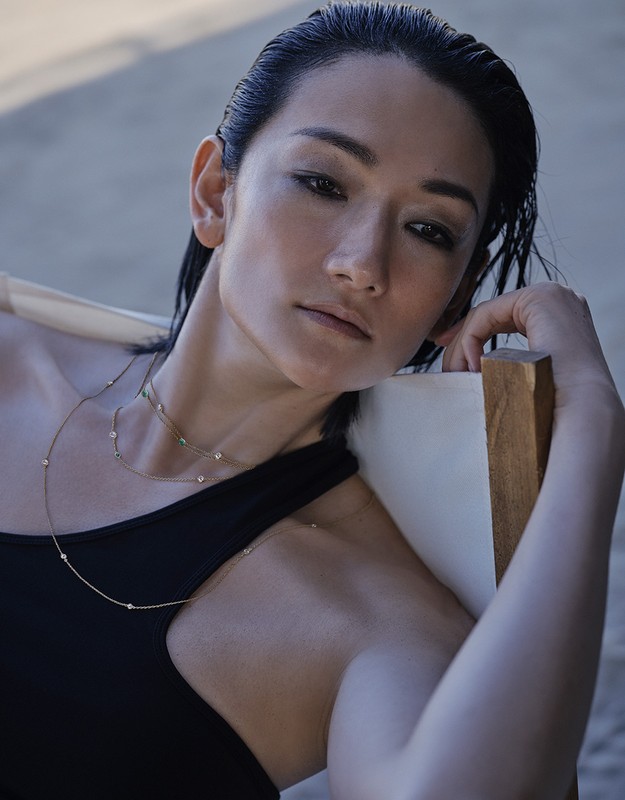 Close-up portrait of a woman with wet dark hair wearing delicate layered gold necklaces and a black top, photographed by Takay.
