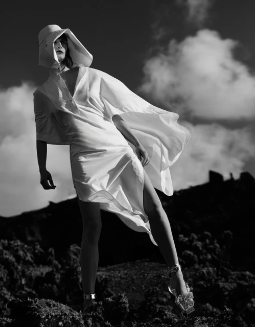 Fashion shot of a woman in a white dress and hat, standing in an outdoor rocky environment under a cloudy sky in black and white.