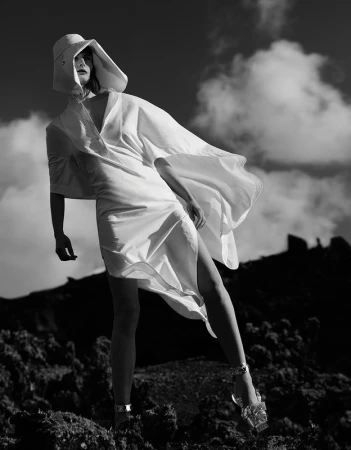 Fashion shot of a woman in a white dress and hat, standing in an outdoor rocky environment under a cloudy sky in black and white.