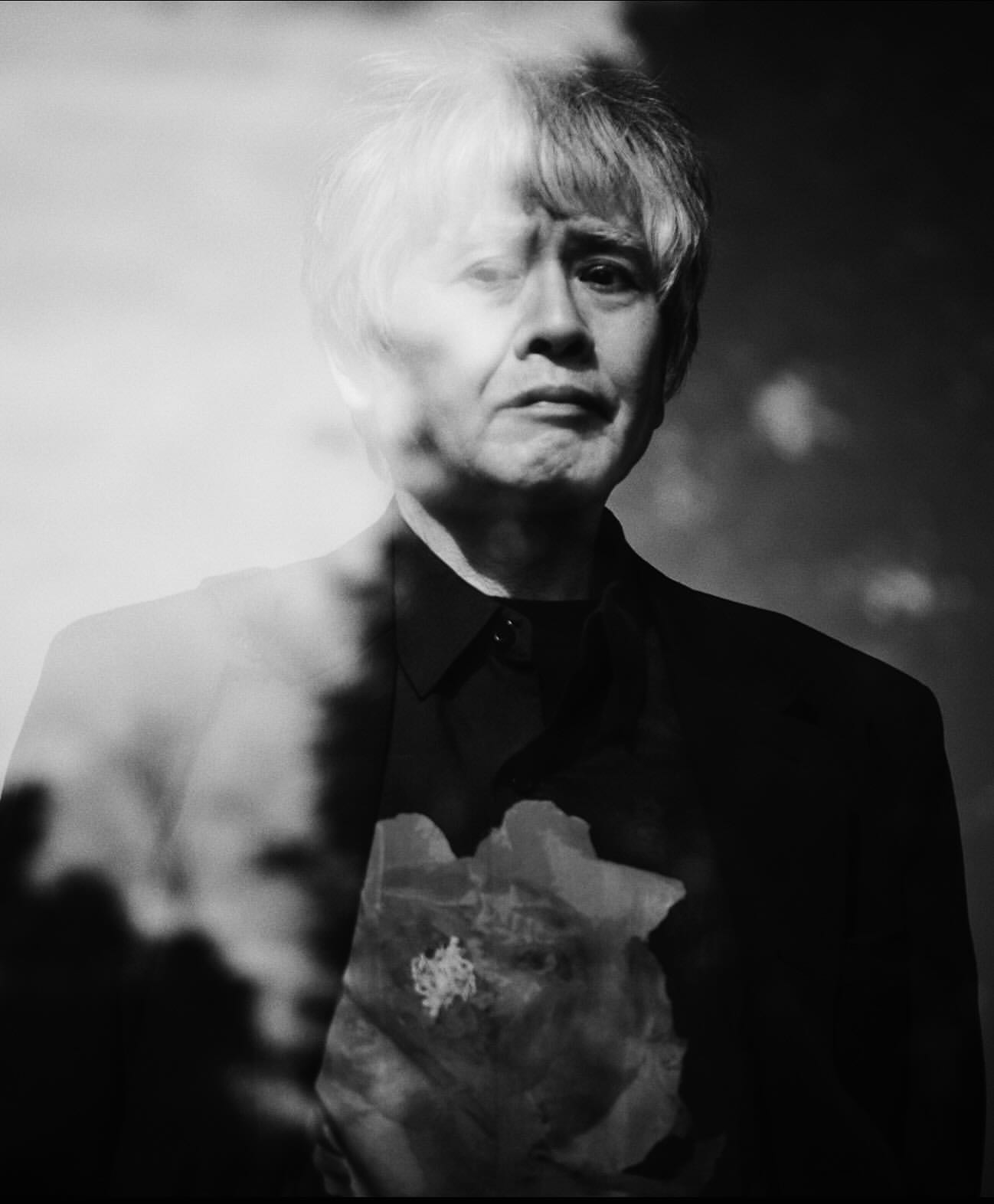 Black-and-white portrait of an adult with short hair in a suit, superimposed with an out-of-focus flower and sky.