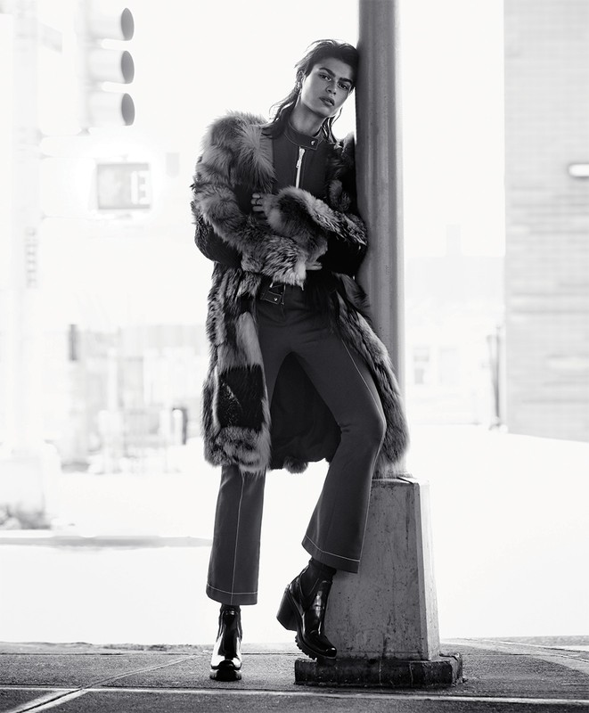 A model leans against a concrete column in a black and white fashion shot, wearing a fur coat, dark pants, and black boots in an urban setting.