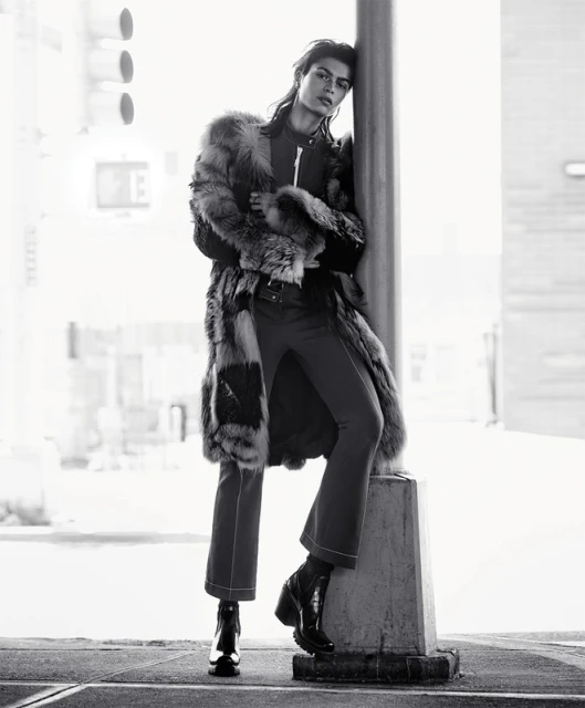 A model leans against a concrete column in a black and white fashion shot, wearing a fur coat, dark pants, and black boots in an urban setting.