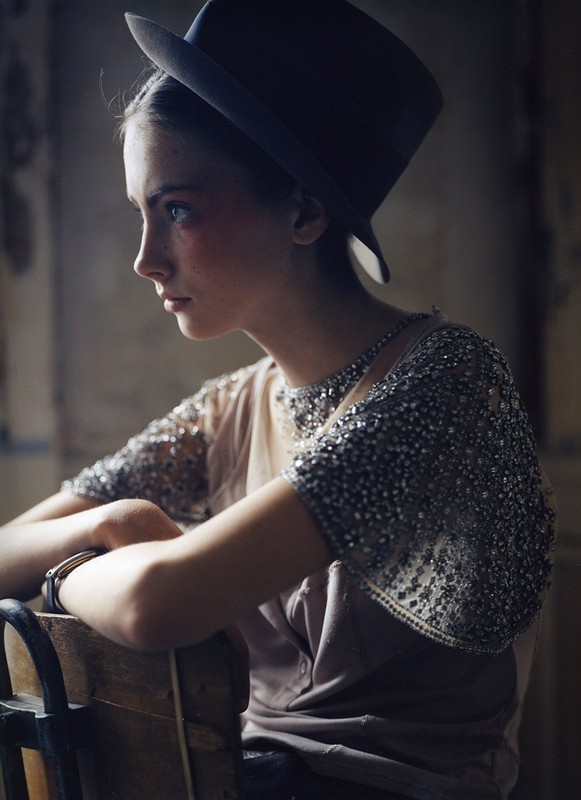 Young woman in profile wearing a black hat and embellished top, seated indoors, showcasing fashion and portrait photography.