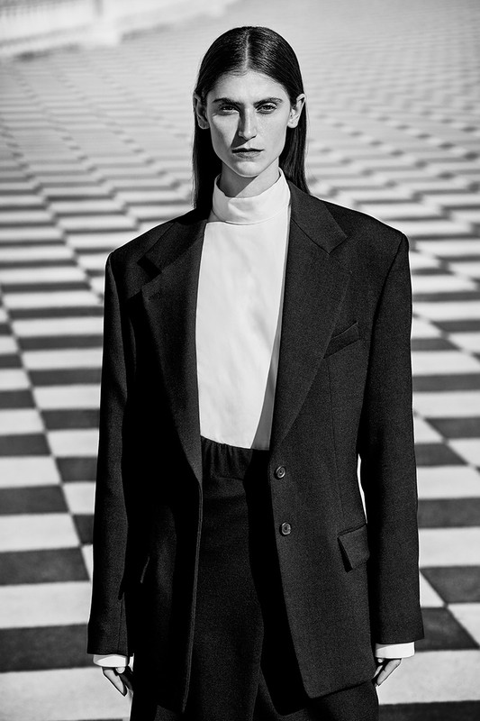 Black and white fashion portrait of a woman wearing an oversized dark blazer and light turtleneck, posing against a checkerboard background.