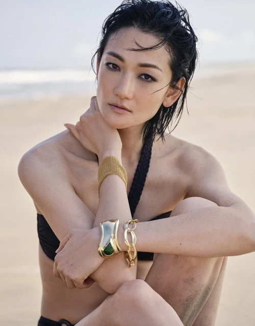 Portrait of an Asian woman on a beach, wearing a black bathing suit and gold bracelets in soft light.