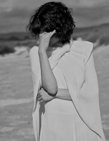 Monochrome portrait of a person with short curly hair covering eyes with a hand, wearing draped white fabric outdoors.